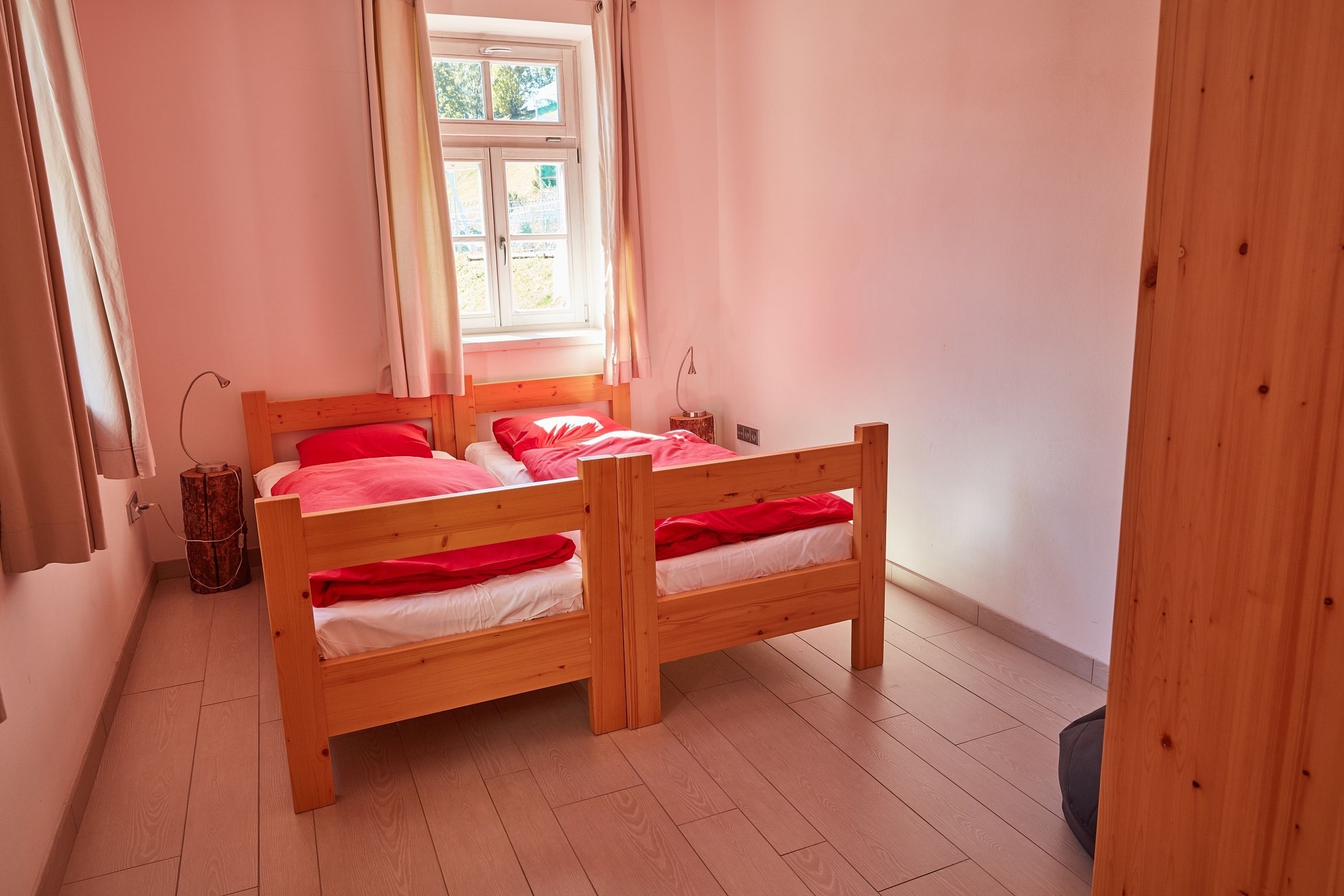 A room with a wooden bunk bed, a window with curtains, a small table with a lamp, and a trash bin.