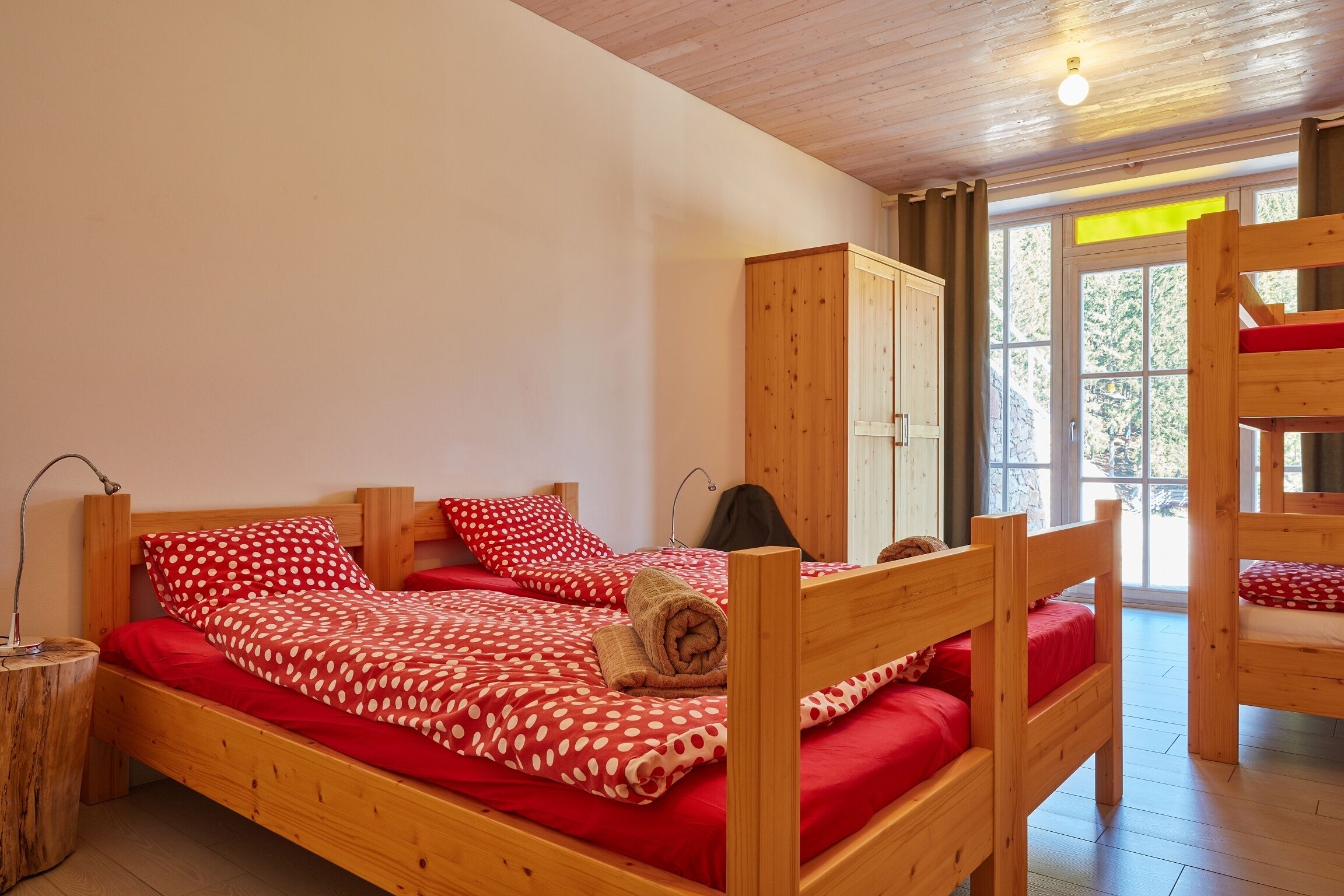 Un lit superposé en bois avec une literie rouge, une armoire en bois et une fenêtre donnant sur des arbres.