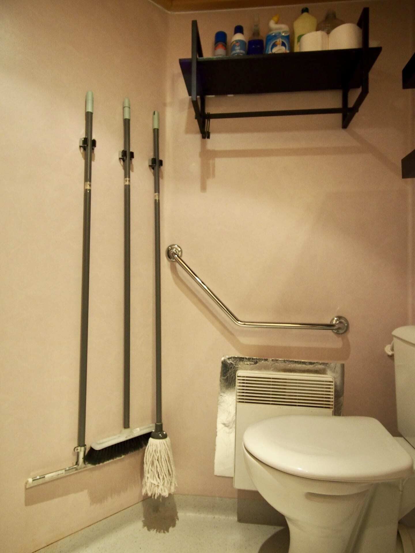 A bathroom with a toilet, a mop, a broom, and a shelf with toiletries.