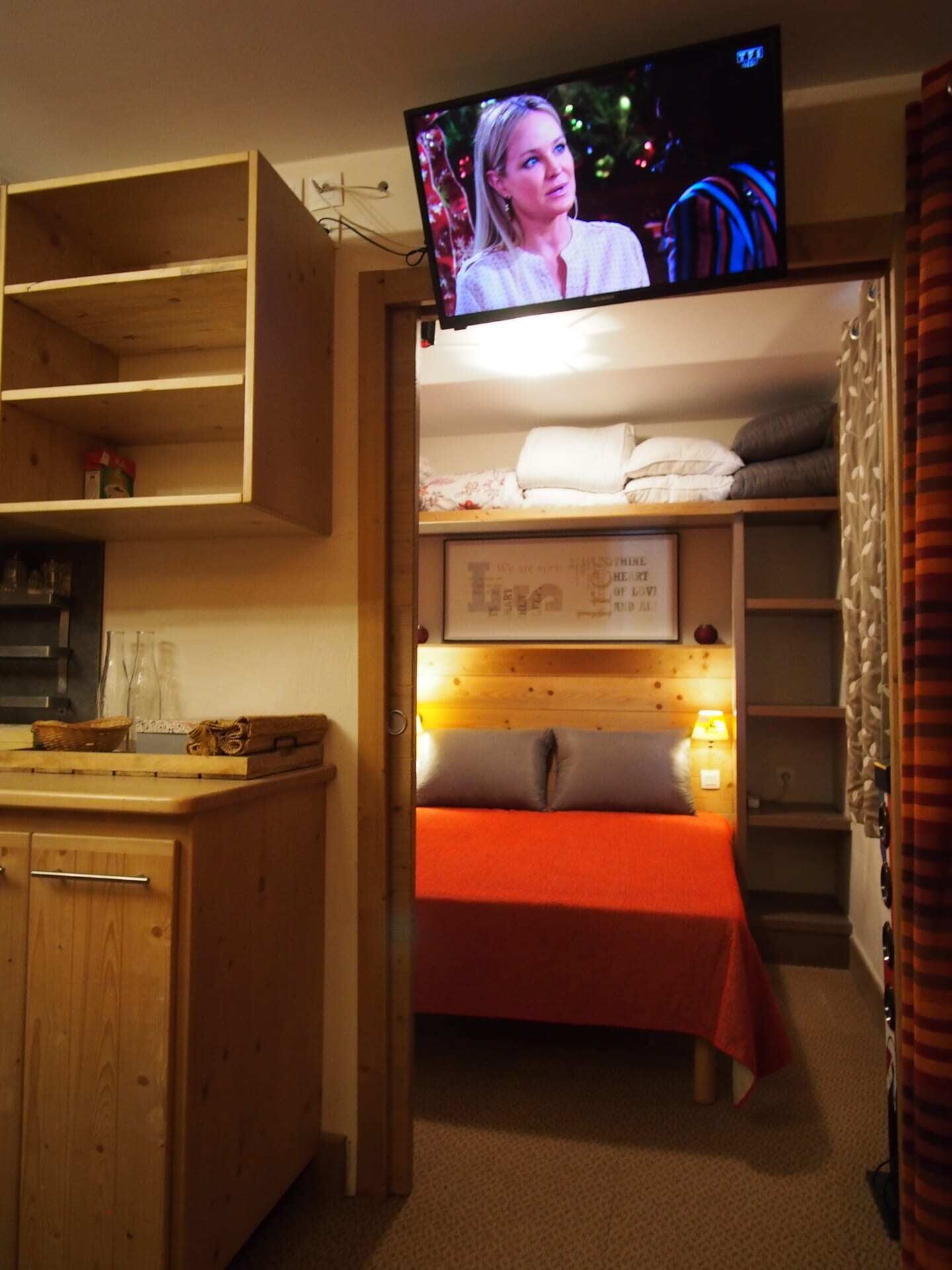 Quarto compacto com cama beliche, pequena cozinha e televisão fixada na parede.