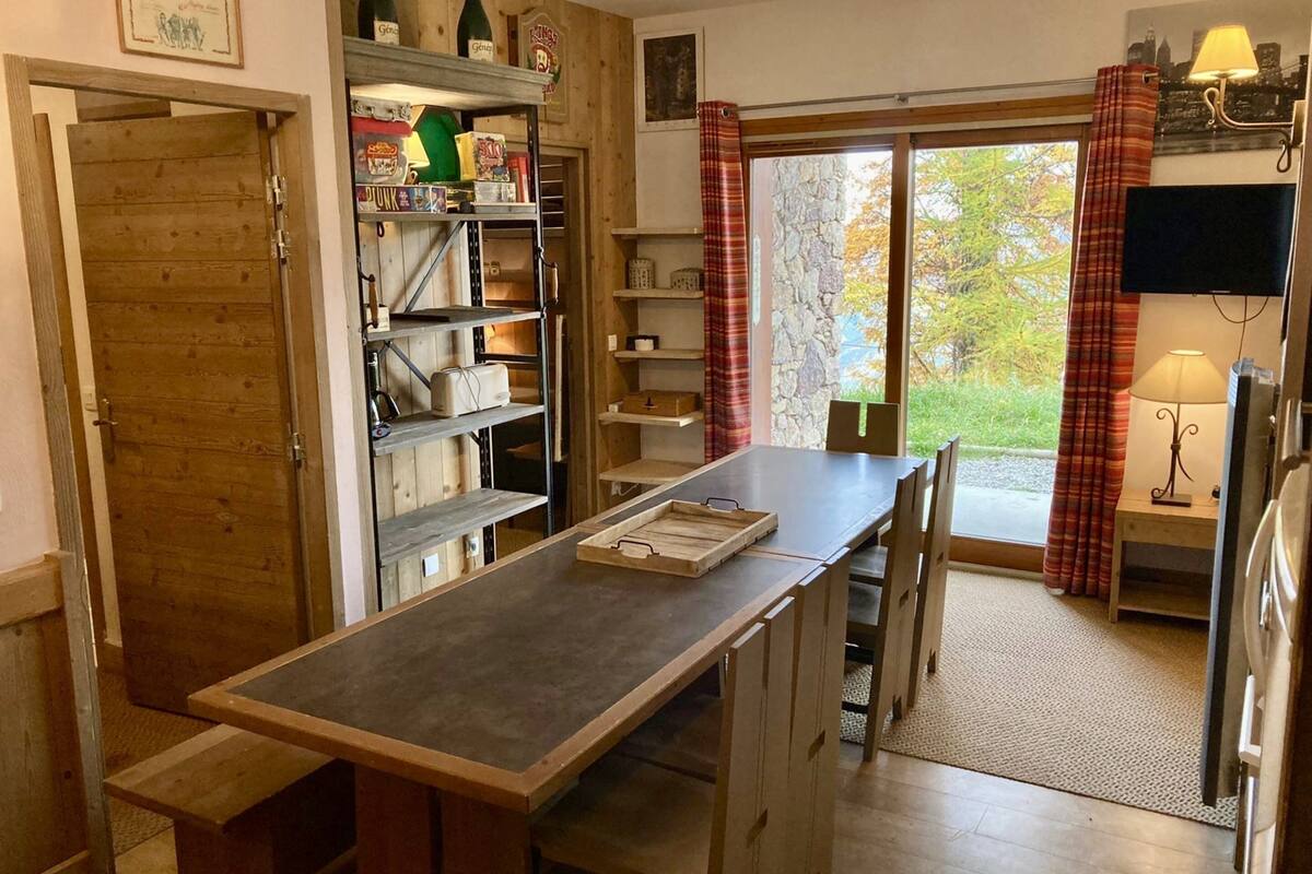 A kitchen with a wooden table, shelves with various items, and a view of the outdoors through a glass door.