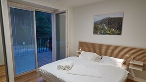 A bedroom with a bed, white linens, a wooden headboard, a nightstand, a wall-mounted lamp, and a painting of a landscape.
