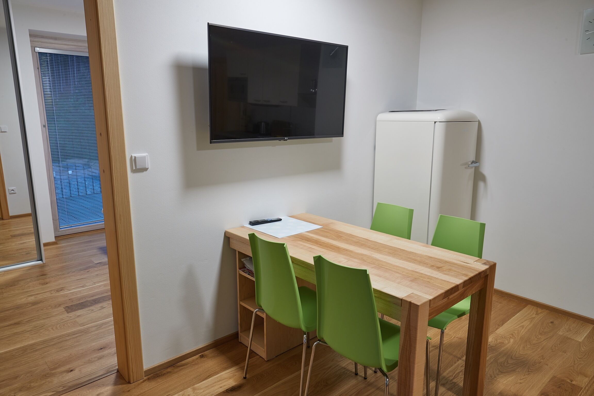 Ein Zimmer mit Holztisch, grünen Stühlen, einem Flachbildfernseher und einem weißen Schrank.