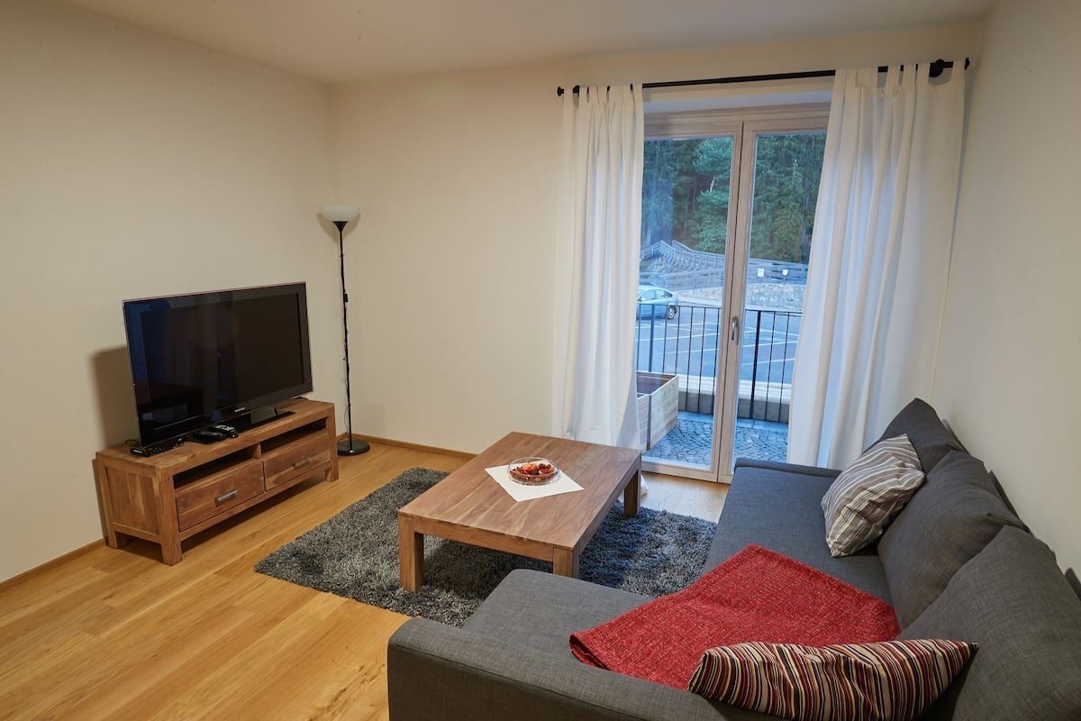 A modern living room with a flat-screen TV on a wooden stand, a small wooden table with a bowl, a grey sofa with striped pillows, and a view of a balcony with a railing.