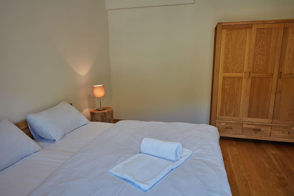 A bedroom with a bed, pillows, a nightstand with a lamp, and a wooden wardrobe.
