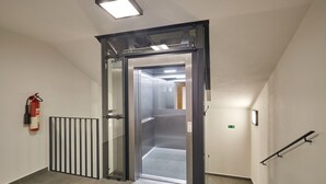 A modern elevator entrance with a glass door, a fire extinguisher on the wall, and a handrail on the right.