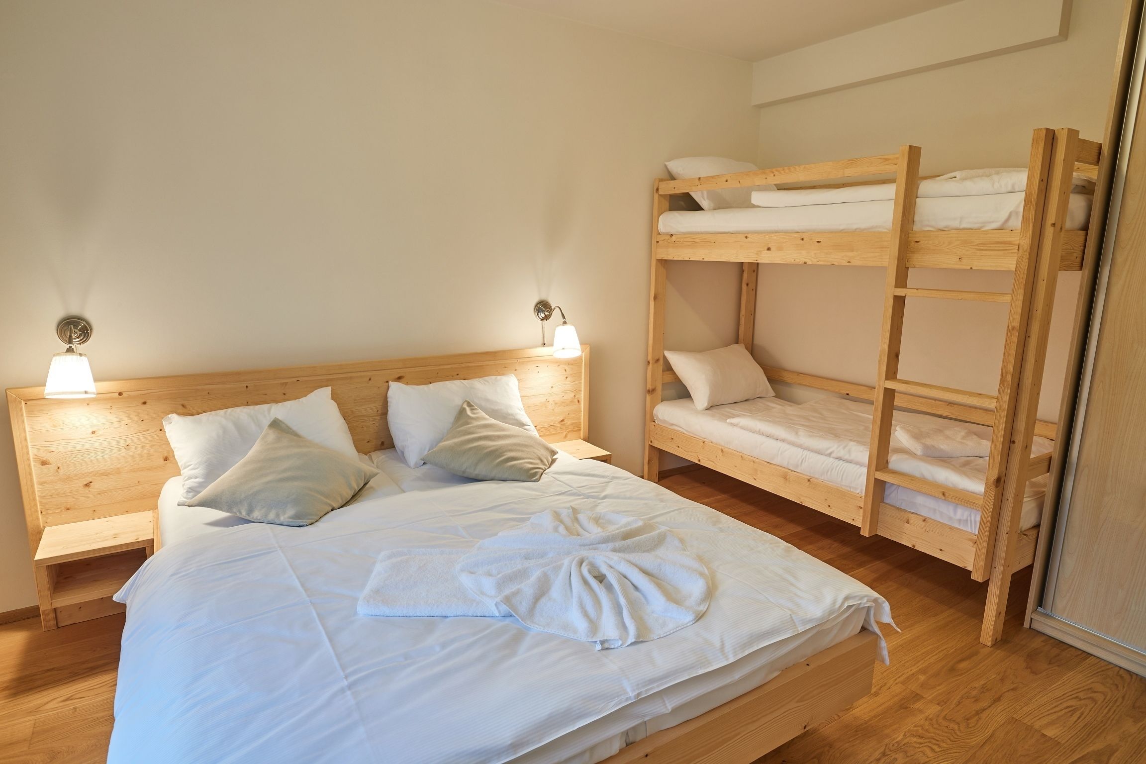 A room with a wooden bunk bed, a single bed with white bedding, and two bedside lamps.