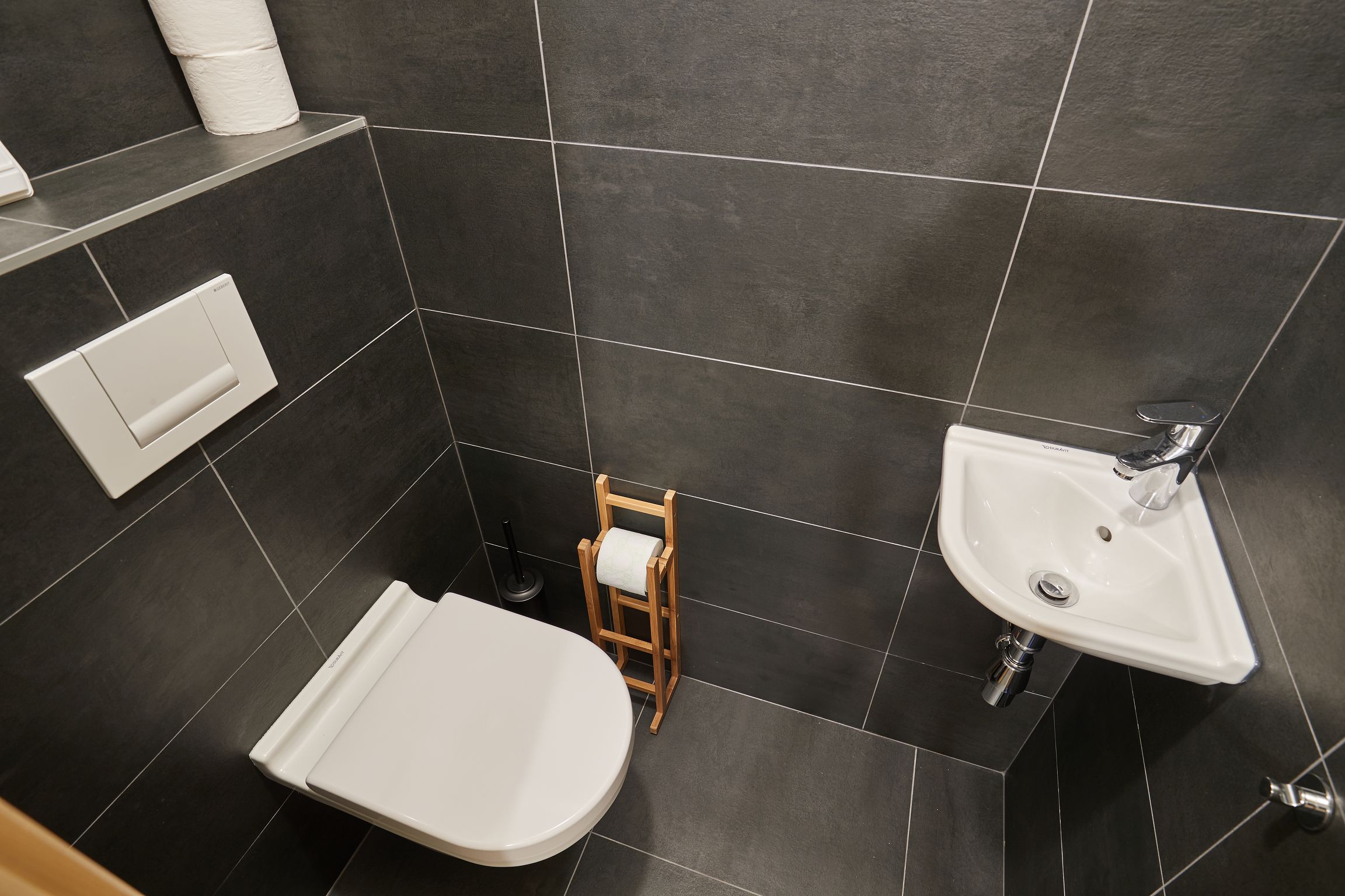 A modern bathroom with a white toilet, a wooden chair, a white sink, and a towel dispenser.