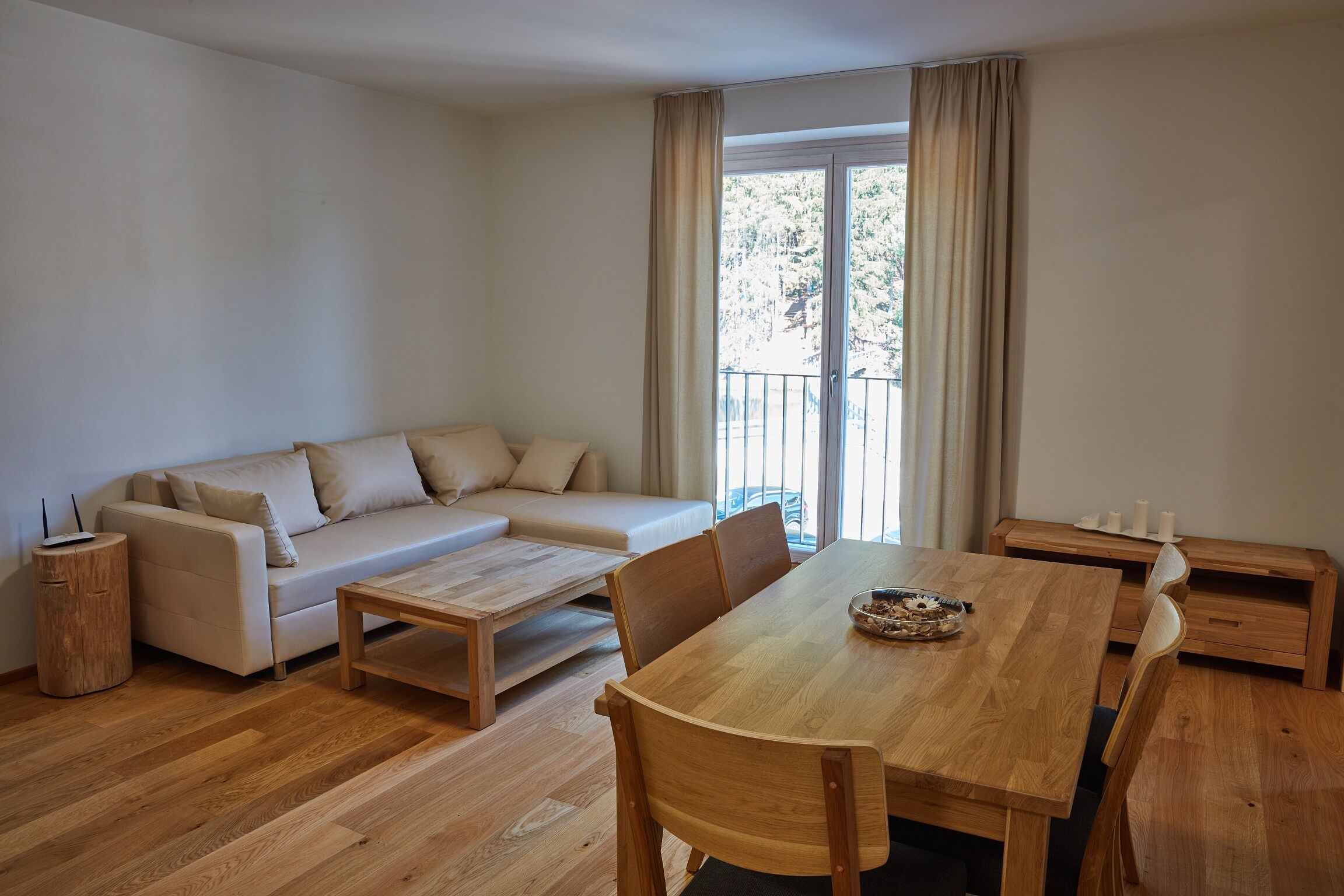 A modern living room with a wooden dining table, beige sofa, and a balcony with curtains.