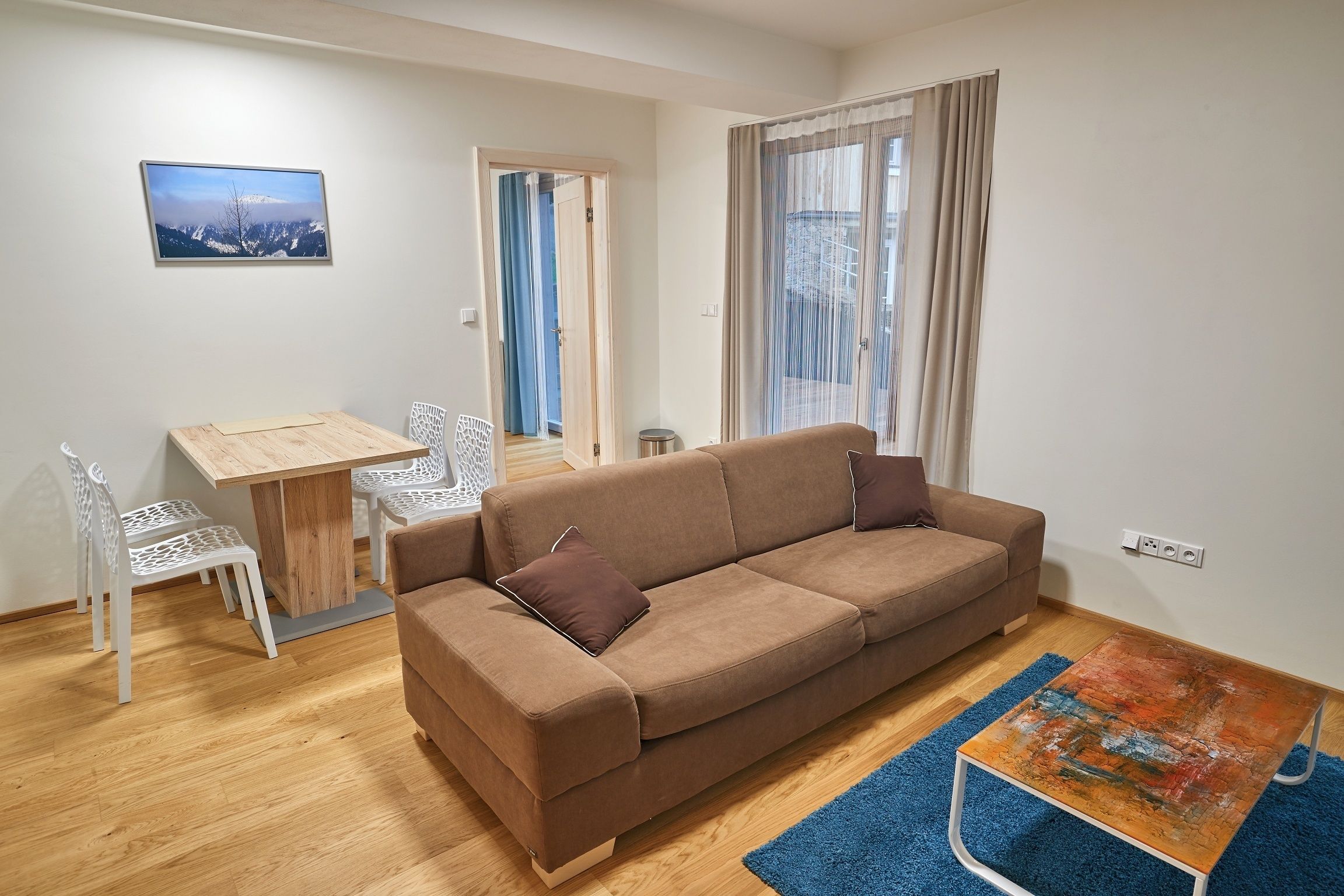 Una sala de estar con un sofá marrón, una mesa de comedor de madera, sillas blancas, un tapete azul y un cuadro en la pared.