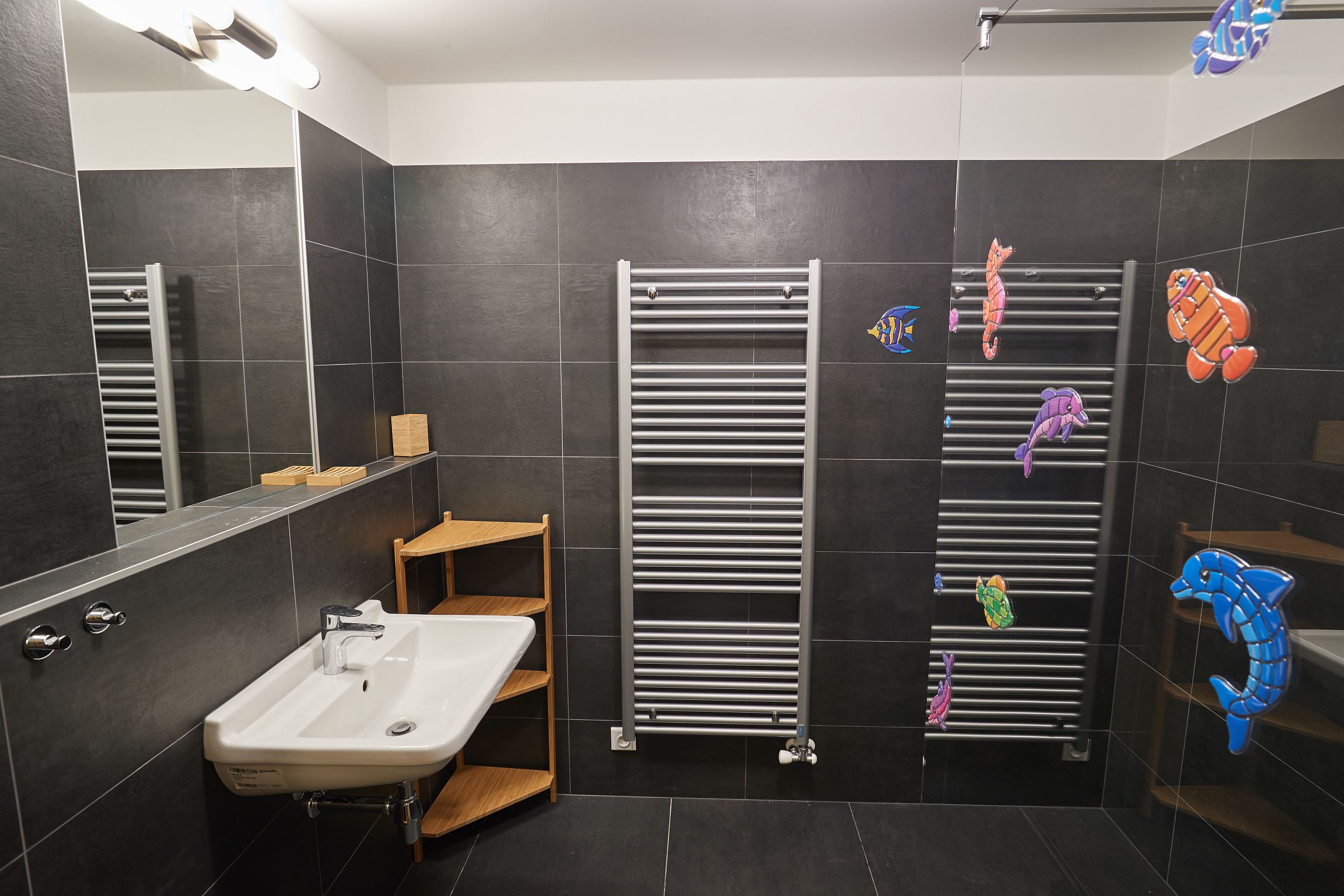 Une salle de bain moderne avec un lavabo blanc, des murs recouverts de carreaux foncés et un porte-serviettes décoré de poissons colorés.