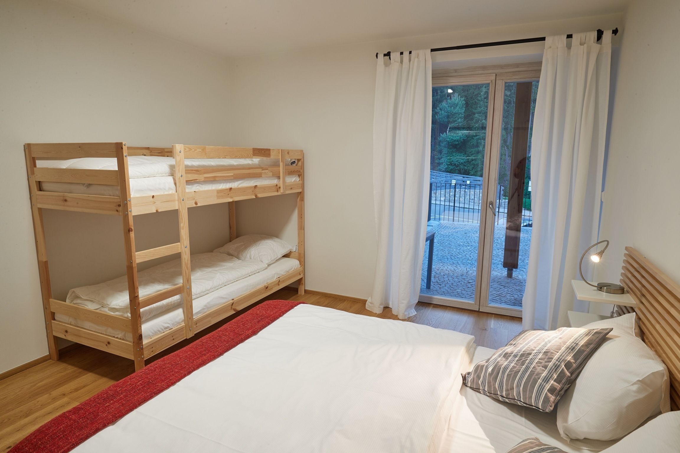 Une chambre avec un lit superposé, un lit simple, un tapis rouge, une lampe et une vue sur l’extérieur grâce à une porte vitrée.