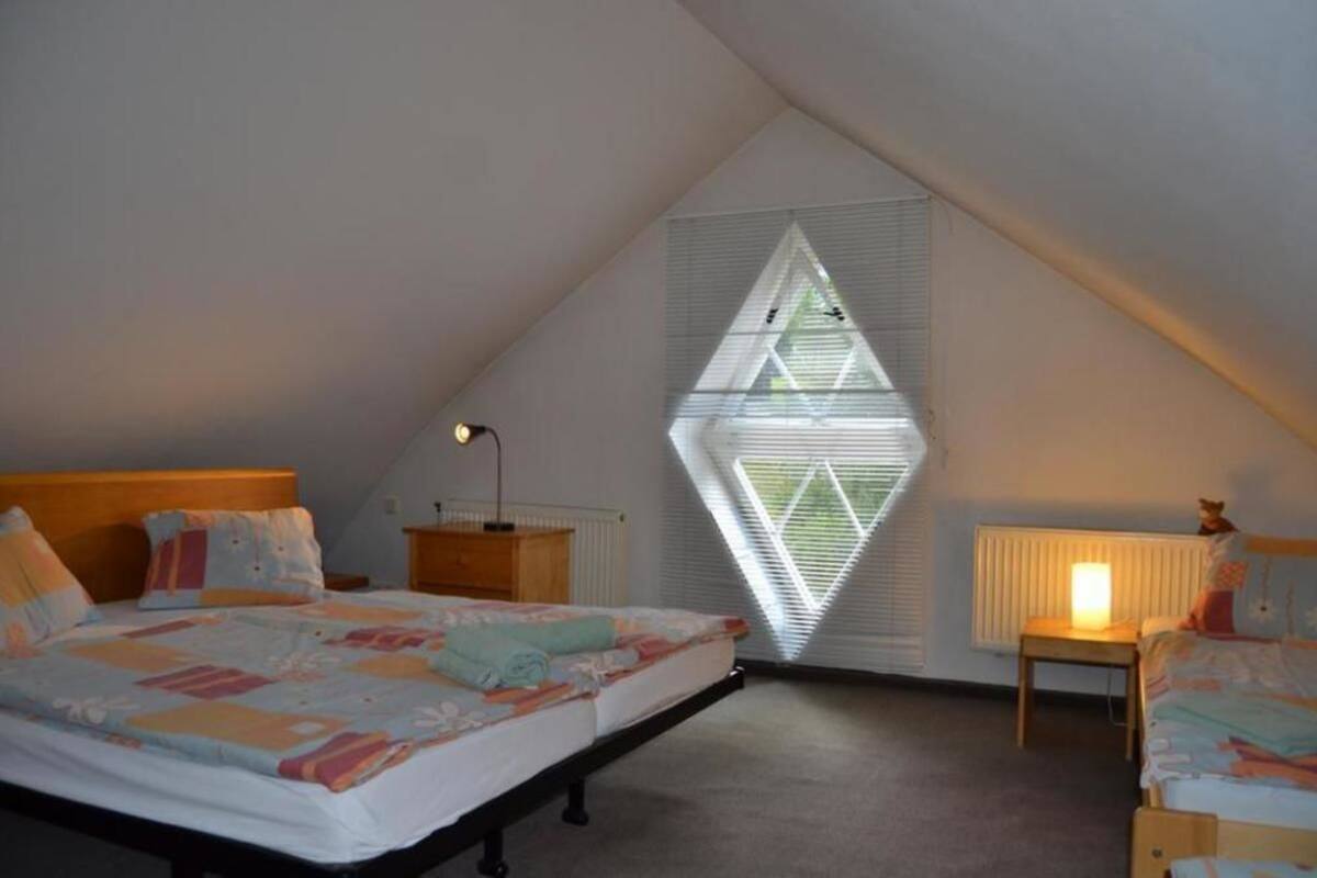 Ein Schlafzimmer mit schrägen Wänden, einem Bett mit farbenfroher Decke, einem Nachttisch mit Lampe und einem Fenster mit Jalousien.