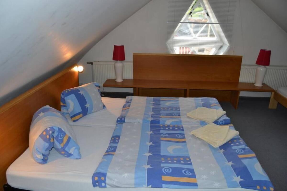 Ein Schlafzimmer mit schrägen Wänden, einem hölzernen Kopfteil, einem Bett mit gemustertem Überwurf und zwei Nachttischen mit Lampen. Ein Fenster mit Rollläden ist ebenfalls zu sehen.