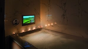 A spa room with a marble wall, a bathtub, and a television displaying a landscape.