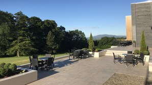 An outdoor patio with tables and chairs, surrounded by greenery and a view of distant hills.