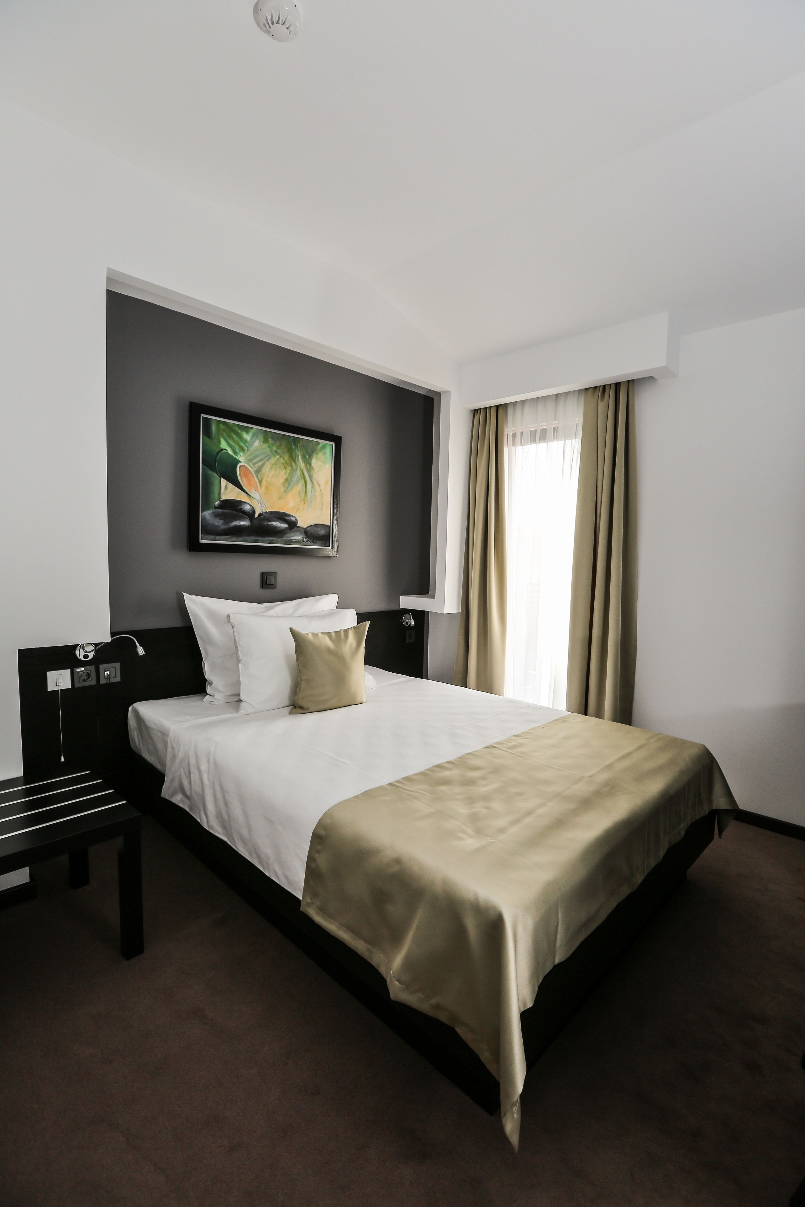 A hotel room with a bed, a bedside table, a headboard, a painting on the wall, and a window with curtains.