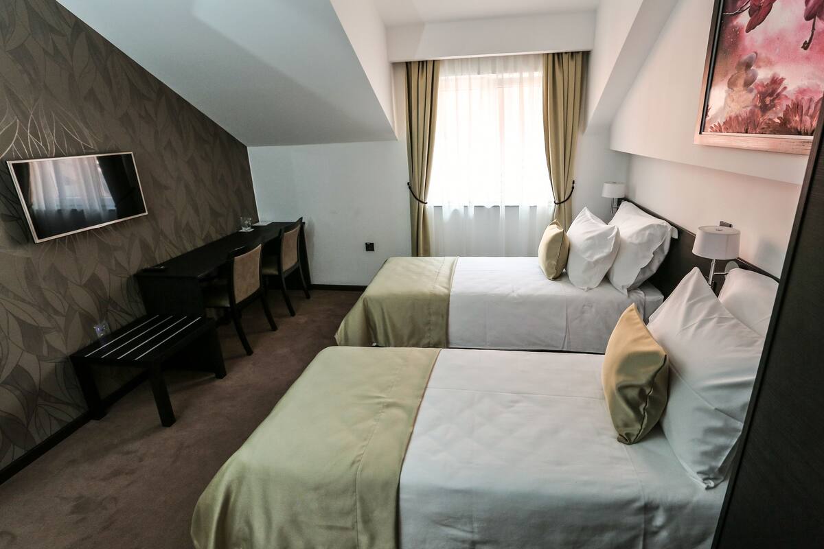 A hotel room with two beds, a desk, a TV, and a window with curtains.