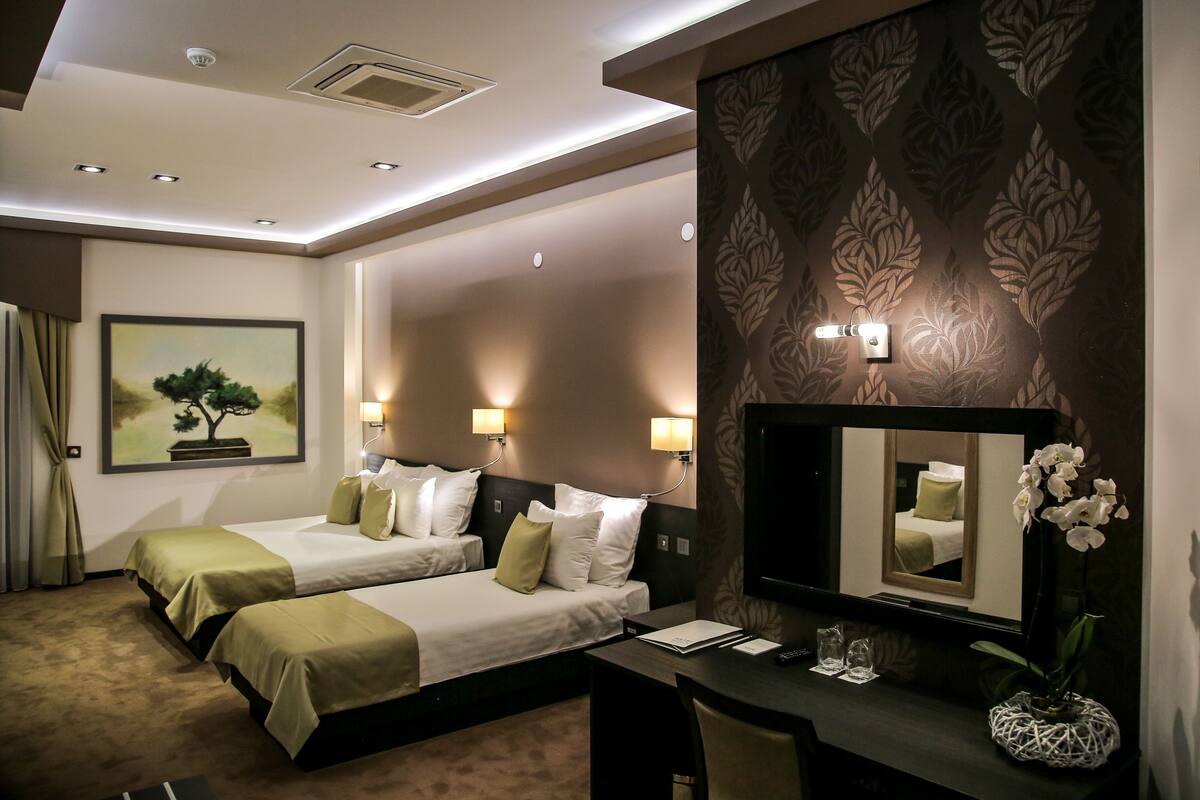 A hotel room with two beds, a desk, a chair, a mirror, and a vase with flowers.