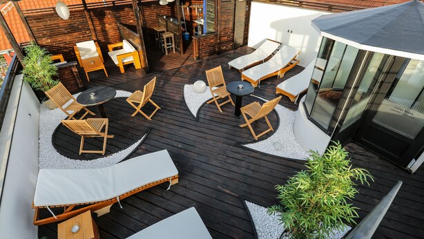 Terraza de madera con sillones y mesas, rodeada de paredes blancas y un cerramiento de vidrio.