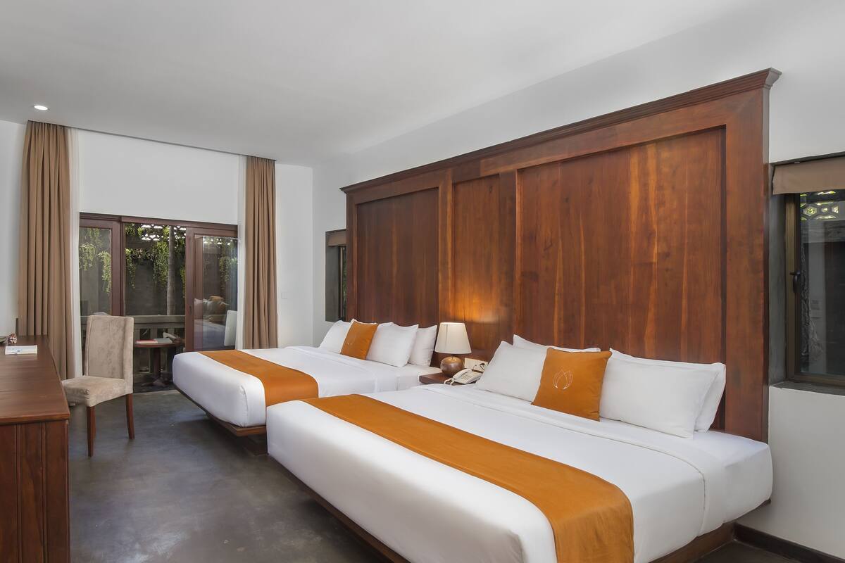 A hotel room with two beds, a desk, a chair, and a view of greenery outside.