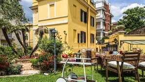 A courtyard with a yellow building, outdoor seating, and a variety of plants and flowers. - Villa Paganini B&B (Rome)