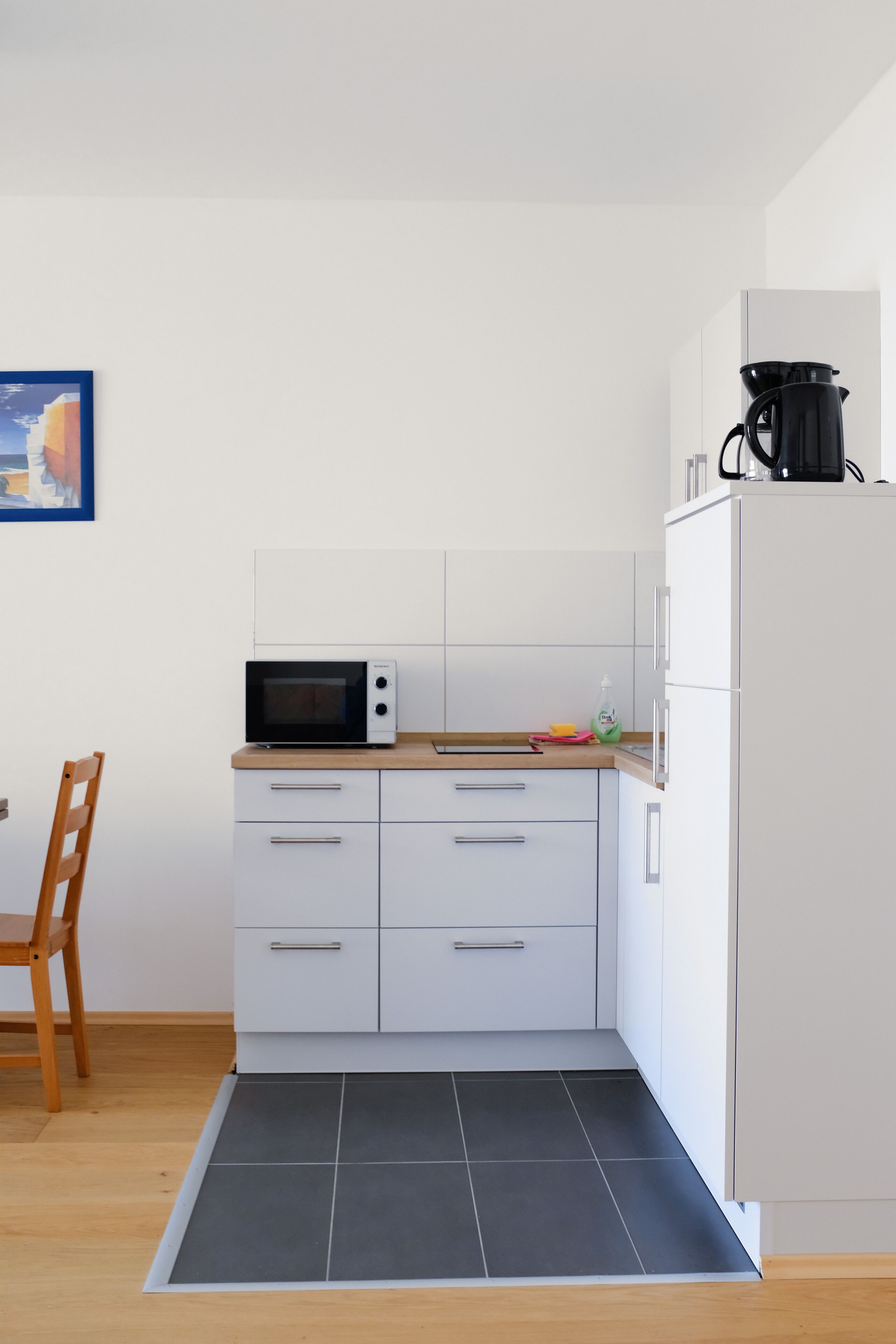 Una cocina compacta con armarios blancos, un microondas, una cafetera negra y un refrigerador.