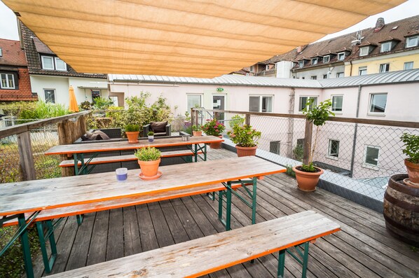 Een houten terras met picknicktafels en plantenbakken, omgeven door een hek van gaas en gebouwen.