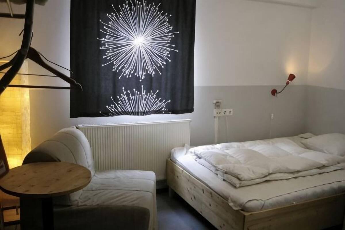 Une petite pièce simple, avec un lit, une chaise, une table et un luminaire fixé au mur.
