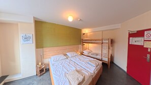 Una habitación con literas, una mesita de noche pequeña con lámpara y una puerta roja con carteles informativos.