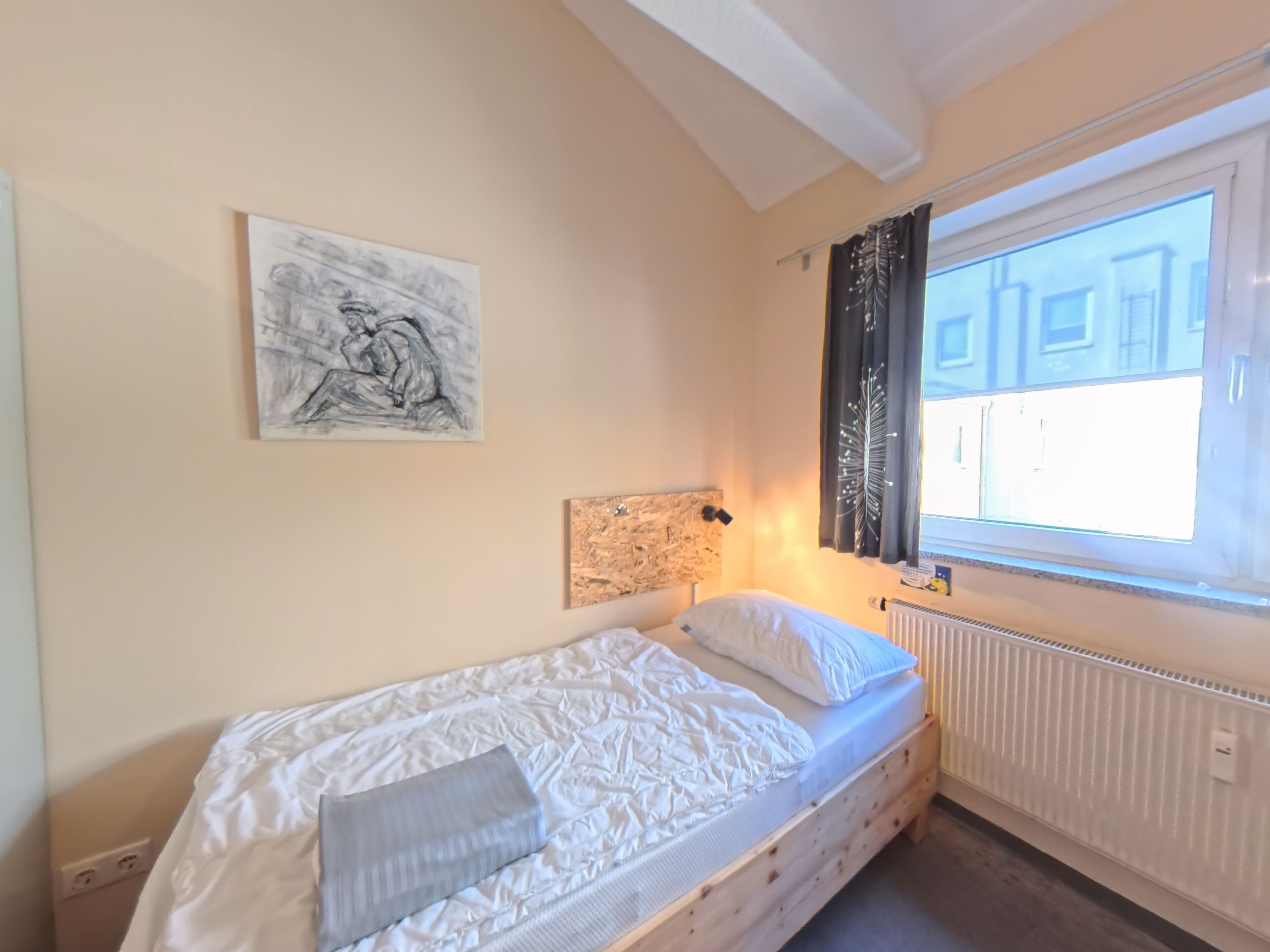 A single bed with a white mattress and pillows, a radiator, a window with curtains, and a wall-mounted artwork.