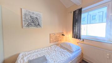 A single bed with a white mattress and pillows, a radiator, a window with curtains, and a wall-mounted artwork.