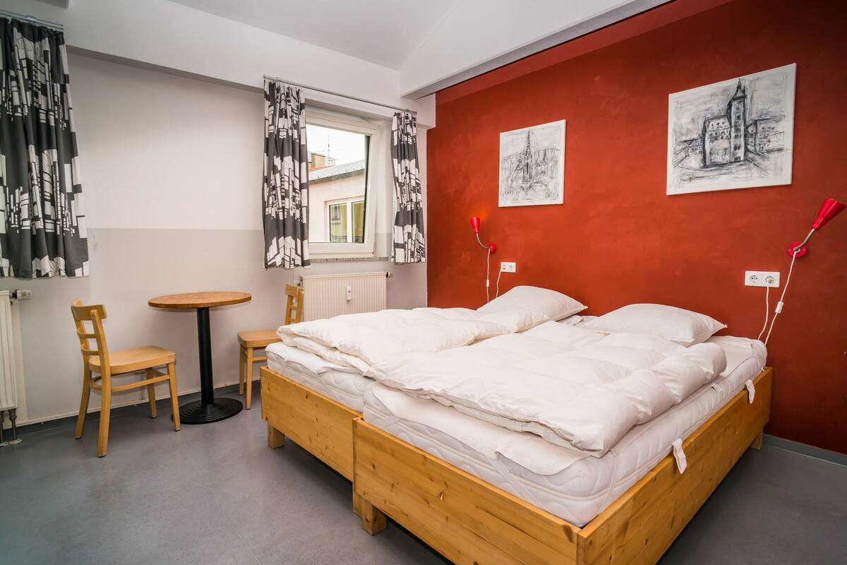 Une chambre à coucher avec un lit en bois, deux chaises, une petite table ronde et une fenêtre avec des rideaux.