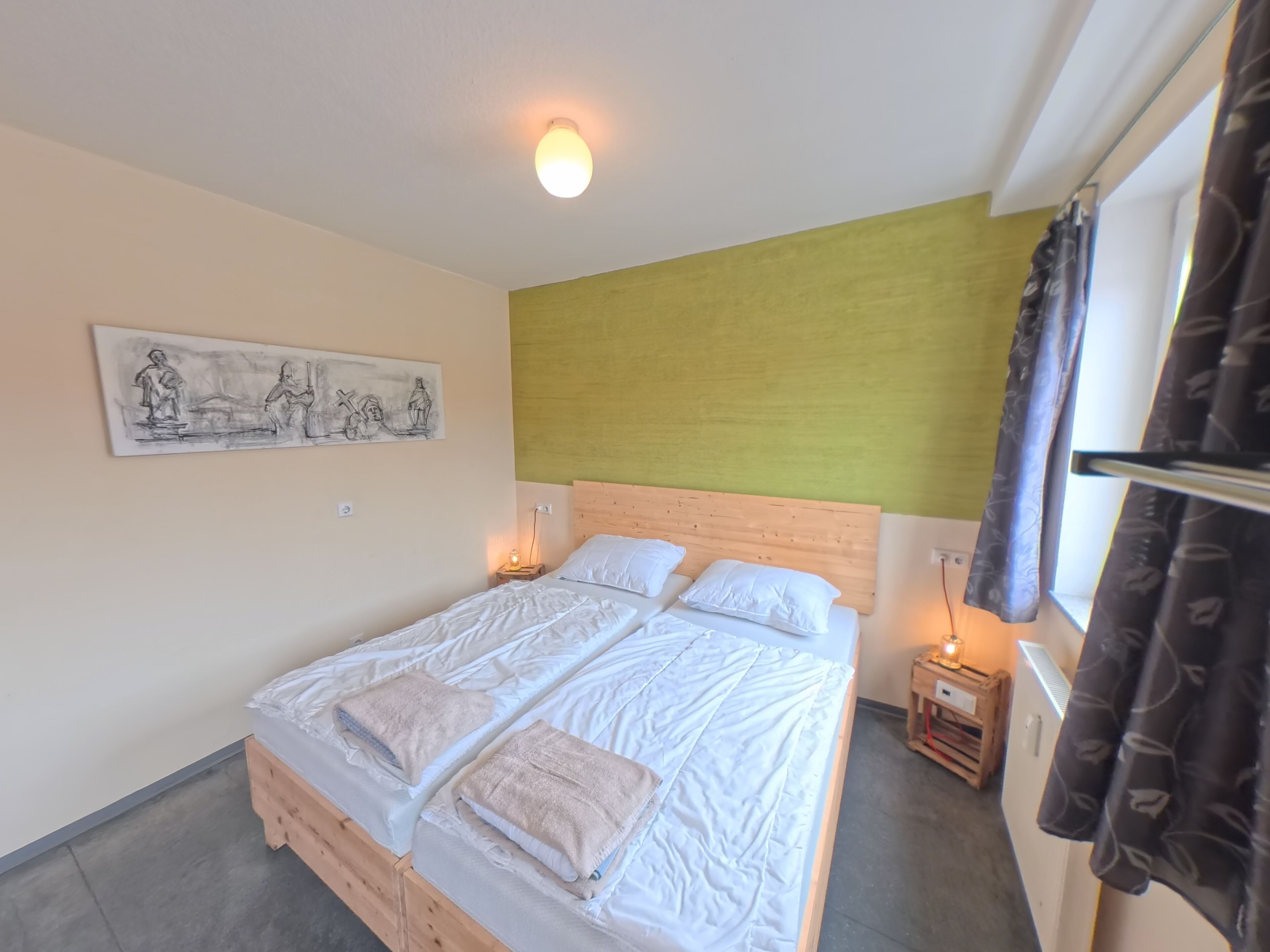 Une chambre à coucher avec un lit en bois, du linge de lit blanc, une tête de lit, une table de chevet, une lampe fixée au mur et une fenêtre avec des rideaux.