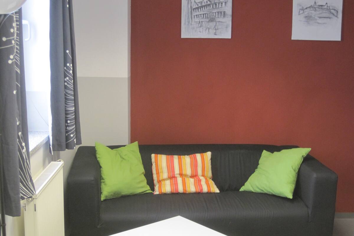 Un salon avec un canapé noir, des coussins à rayures vertes et oranges, et deux œuvres d’art accrochées au mur représentant des bâtiments.