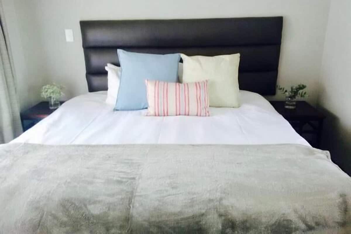 Una cama bien tendida con un cabecero oscuro, ropa de cama blanca y tres almohadas en diferentes colores y diseños.