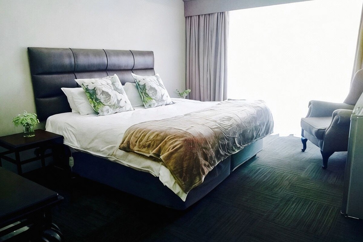 A hotel room with a large bed, a chair, a small table, and a window with curtains.