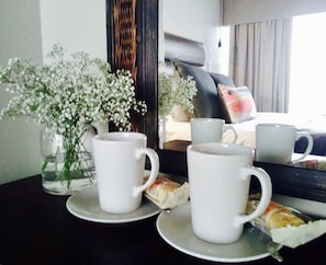 A table with two white cups and saucers, a vase with white flowers, and a window with a view of a building and a parked car. - Shawu Lodge & Conference (Bloemfontein)