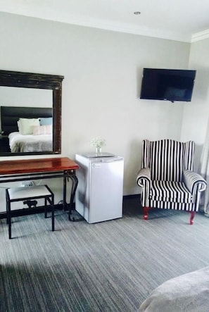 A hotel room with a desk, a small refrigerator, a striped armchair, and a television mounted on the wall. - Shawu Lodge & Conference (Bloemfontein)