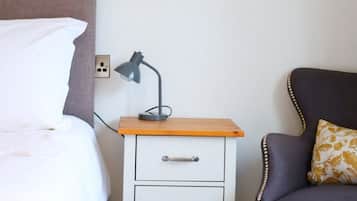 Una camera d'albergo con un letto, un comodino con una lampada, una sedia e un piccolo mobile in legno con cassetti.