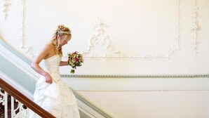 A bride in a white dress descending a staircase with ornate wall decorations. - The Eagle House Hotel (Launceston)