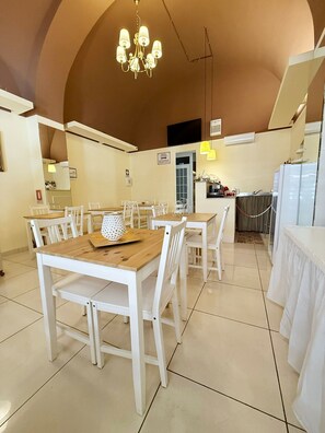 Un comedor con mesas y sillas blancas, una araña y una cocina al fondo.
