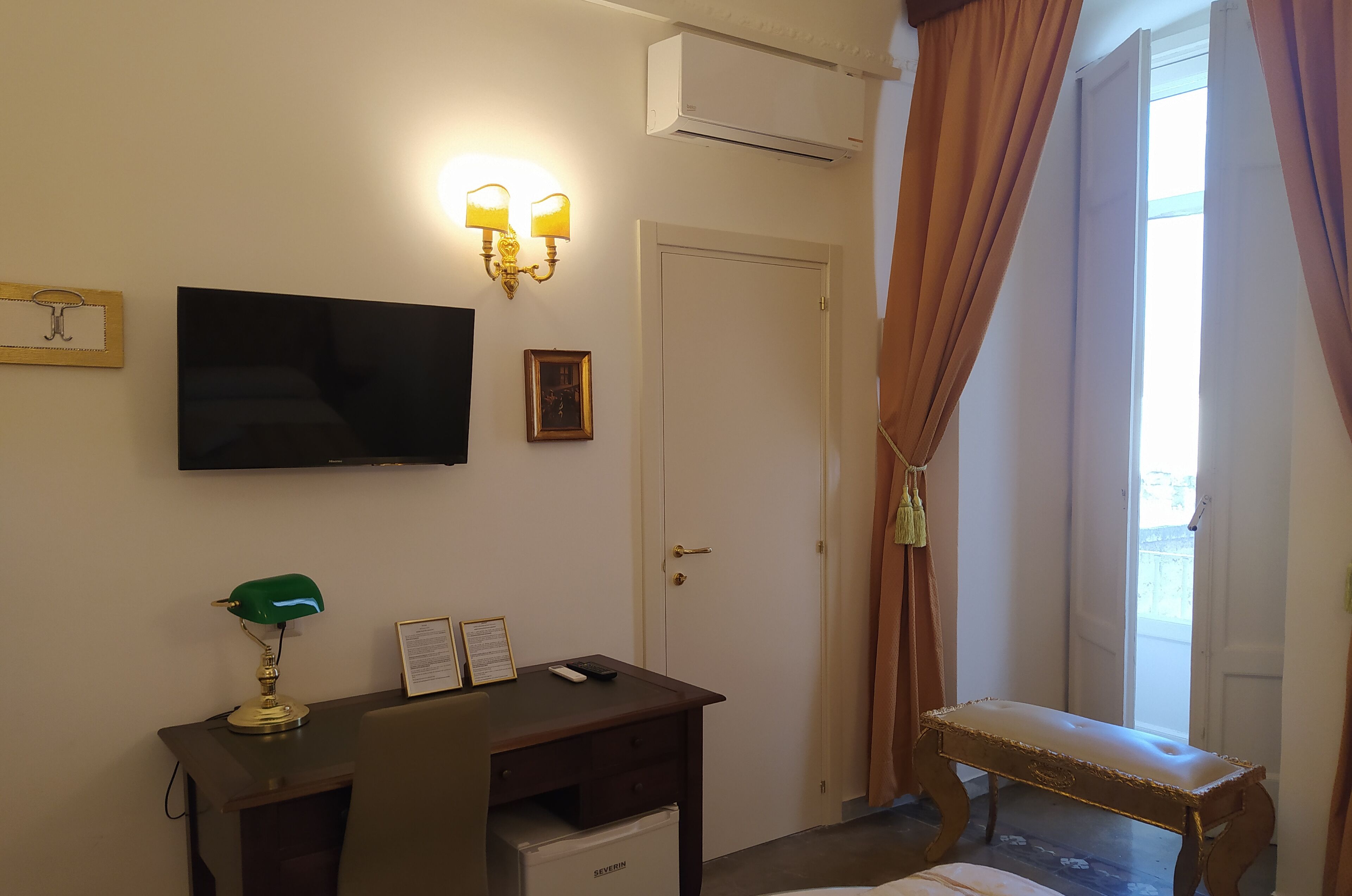 Een hotelkamer met een bureau, een flatscreen televisie, een wandairconditioner en een raam met gordijnen.