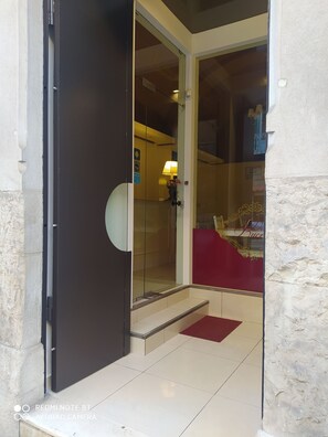 Entrada de hotel con puerta de cristal, alfombra roja y una lámpara en el interior.