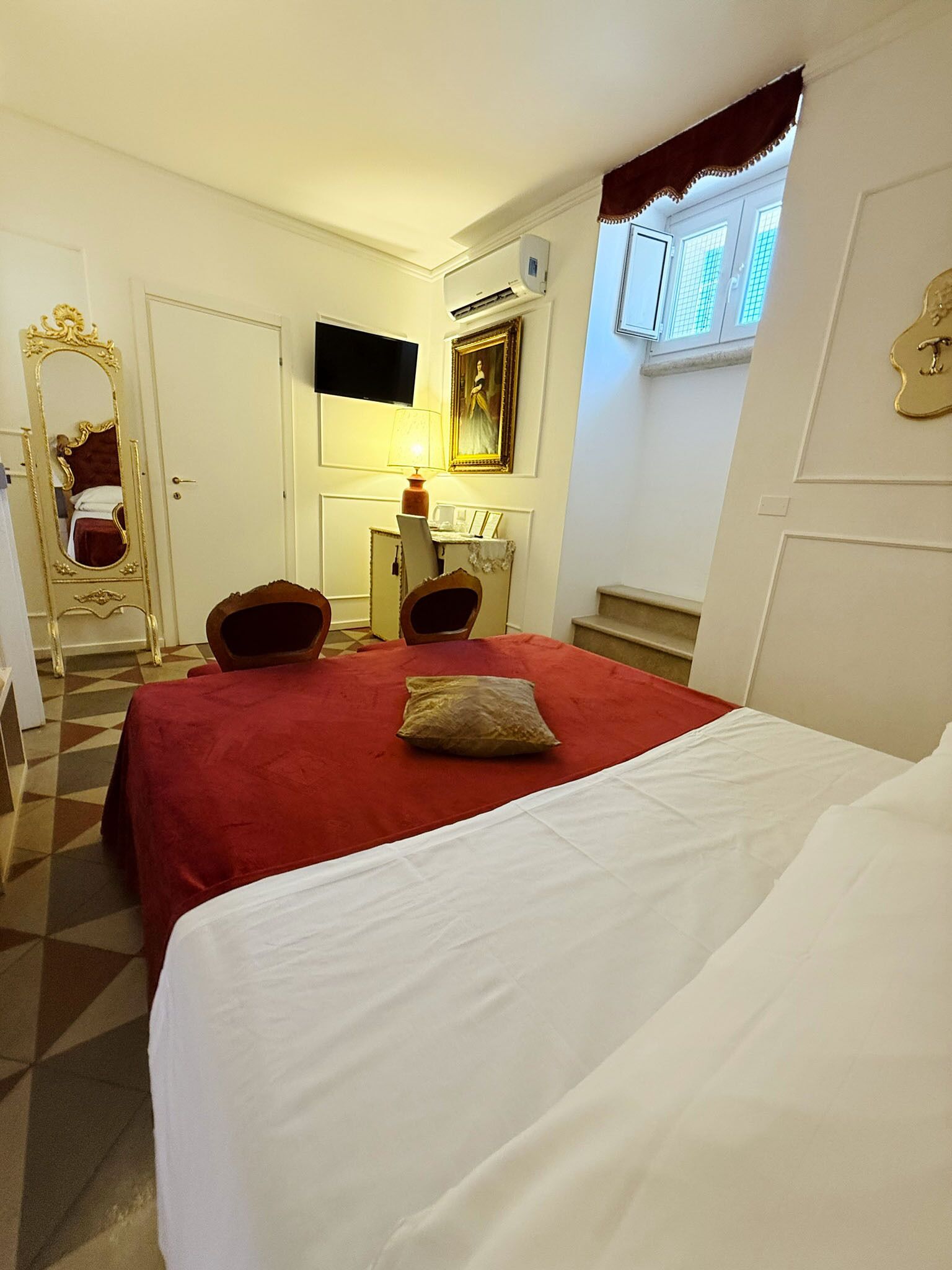 Habitación de hotel con una cama grande, un escritorio con lámpara, una silla, un espejo y un televisor en la pared.