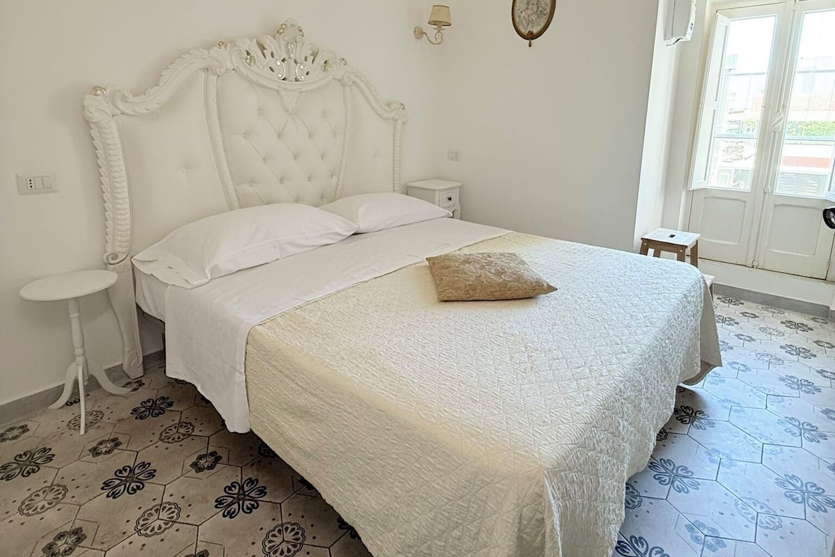 Une chambre à coucher avec un grand lit, une table de chevet, une lampe fixée au mur, une petite table et une fenêtre avec des rideaux.
