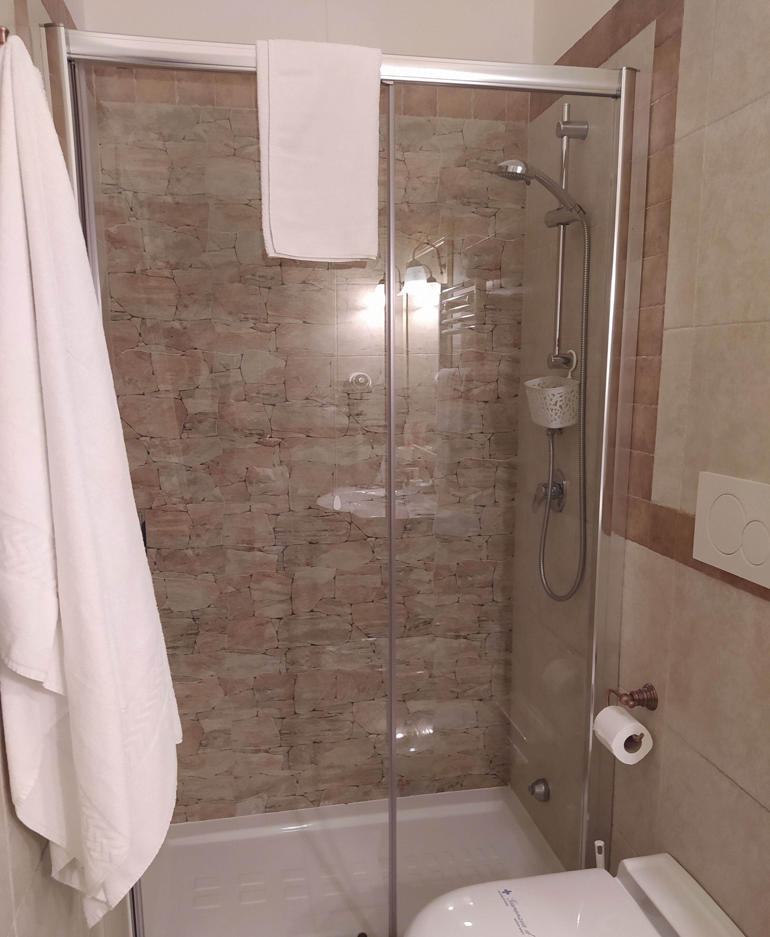 A glass-enclosed shower stall with a stone wall backdrop, a white towel hanging on the door, and a toilet paper holder on the wall.