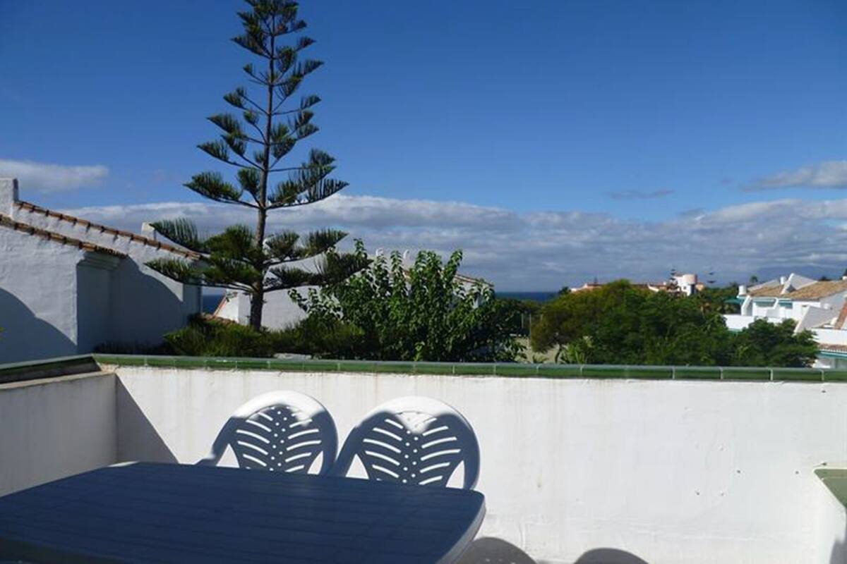 A rooftop terrace with a table and chairs, a pine tree, and a view of buildings and greenery.
