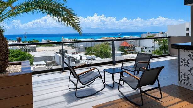 Un balcón con dos sillas y una mesa, con vista al mar y edificios, y una palmera.