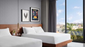 Una habitación de hotel moderna con dos camas, un ventanal amplio con vistas a la ciudad y dos obras de arte enmarcadas en la pared.